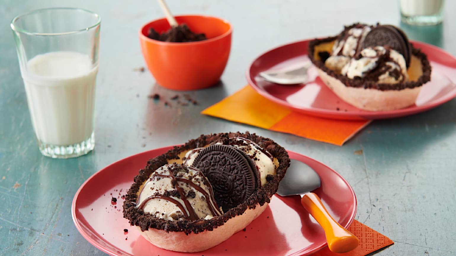 Old El Paso soft tortilla bowls filled with ice cream and Oreos on red plates with spoons, napkins, extra bowl of oreo crumbles and a glass of milk.